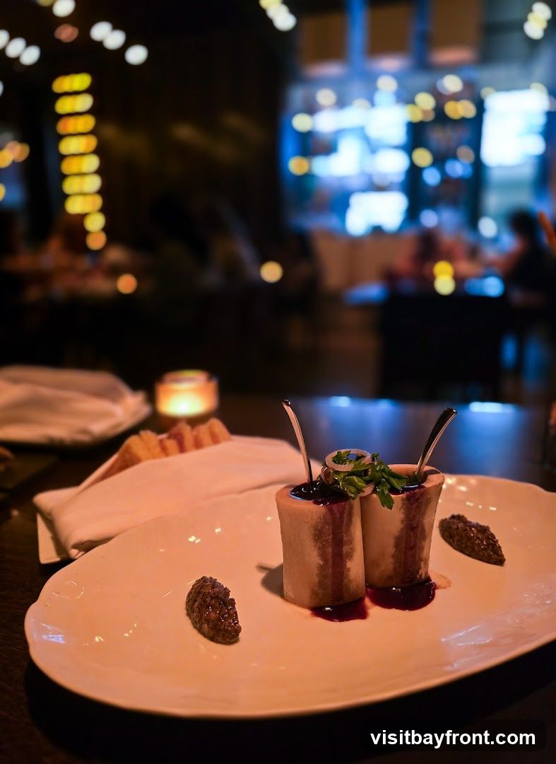 A close-up shot of a gourmet appetizer featuring two pieces of bone marrow, served upright on a white oval plate. The marrow is topped with a dark sauce and garnished with fresh parsley and a ring of thinly sliced onion. Two dollops of a dark, textured relish or spread are placed on either side of the marrow. In the background, a white napkin-wrapped bread basket and a lit candle are visible, along with the blurred ambiance of a dimly lit restaurant interior with patrons and decorative lighting.