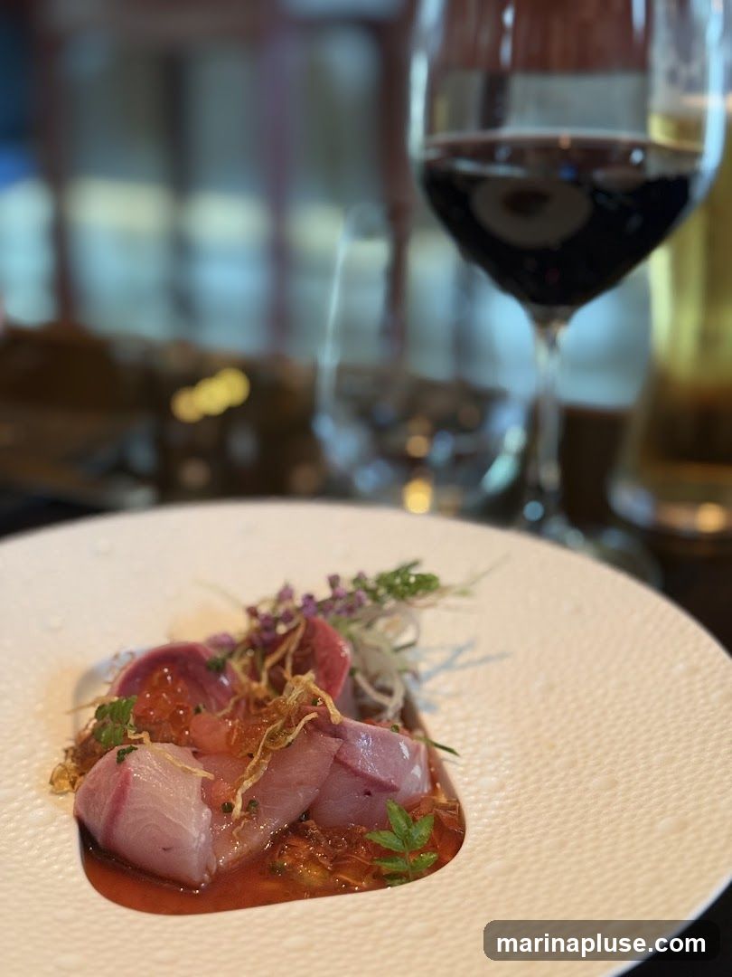 A close-up shot of a delicate seafood appetizer served on a textured white plate. The dish features thinly sliced raw fish, possibly tuna or hamachi, arranged over a bed of finely shredded vegetables and a vibrant red sauce. Garnishes include microgreens and edible flowers. In the background, a glass of red wine is prominently displayed, along with other blurred elements of a restaurant setting.