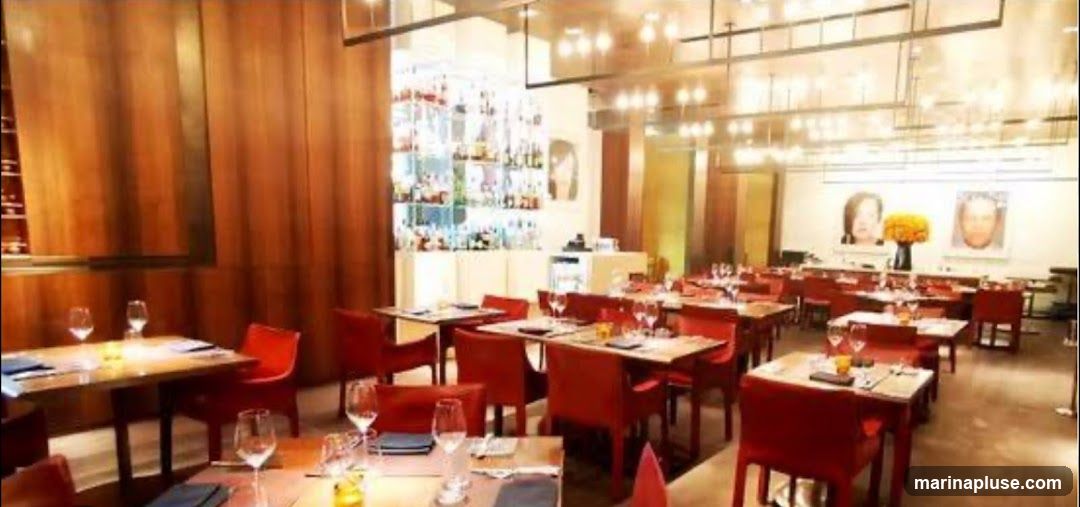 A well-lit restaurant interior with multiple tables set for dining. The tables are covered with white tablecloths and set with glassware and napkins. Red chairs surround the tables. The walls are decorated with portraits and a large floral arrangement. A bar area with shelves stocked with bottles is visible in the background.