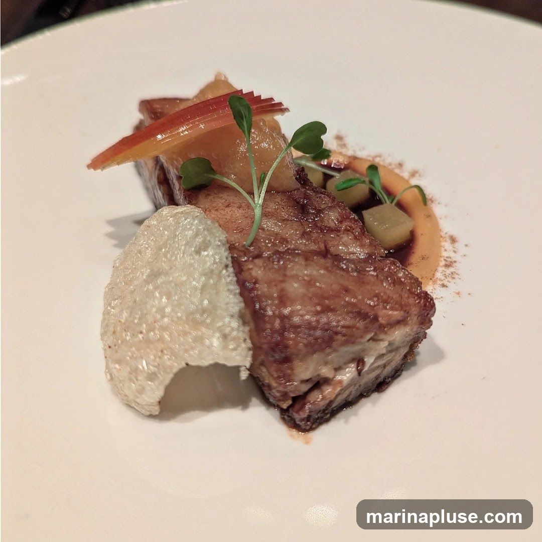 A close-up shot of a gourmet dish featuring a piece of pork belly, garnished with thinly sliced red apple, microgreens, and a crispy cracker. The pork belly is served with a smooth puree and diced apples, dusted with a spice.