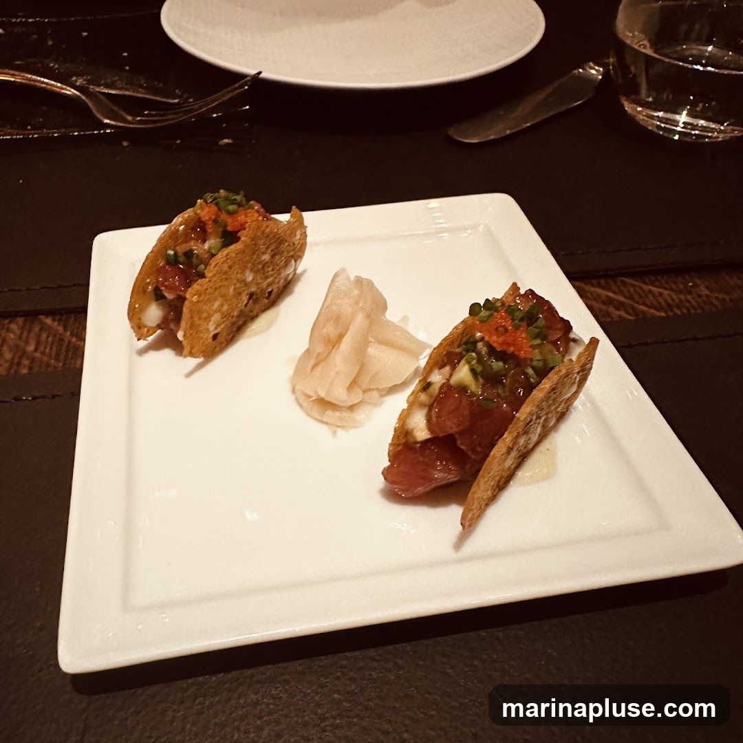 Two tuna tacos served on a white rectangular plate with a side of pickled ginger. The tacos are filled with diced tuna, avocado, and topped with roe and chives. A glass of water and cutlery are visible in the background.