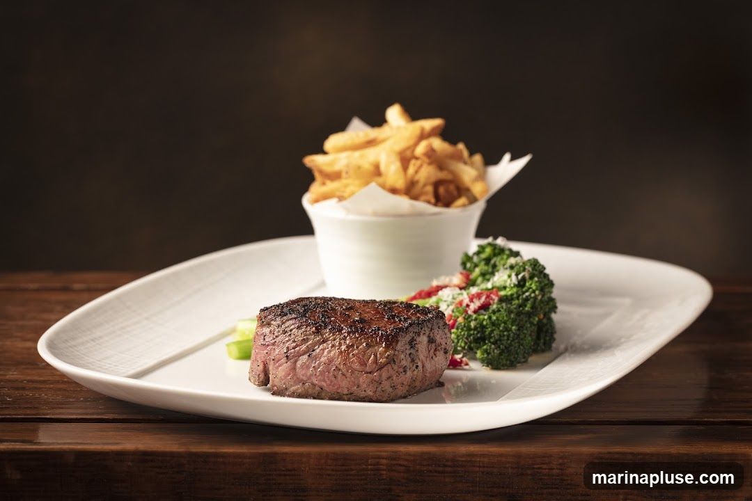 A close-up shot of a steak, french fries, and broccoli served on a white plate. The steak is grilled and appears to be a filet mignon. The french fries are golden brown and served in a white ramekin. The broccoli is steamed and topped with what appears to be parmesan cheese and sun-dried tomatoes. The plate is set on a wooden table with a dark, blurred background.