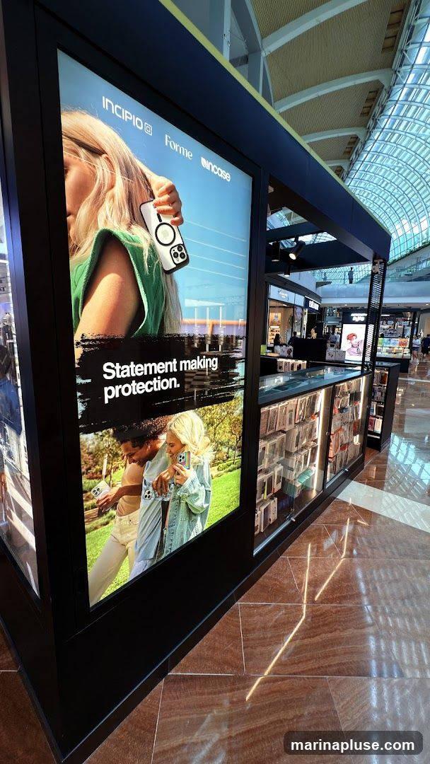 A kiosk in a shopping mall displays phone cases. The kiosk has large advertisements on its sides featuring people holding phones with cases. The text on the advertisement reads "Statement making protection." The kiosk itself is black and has glass display cases filled with various phone cases. The floor is made of polished brown tiles that reflect the light. In the background, other shops and shoppers are visible.