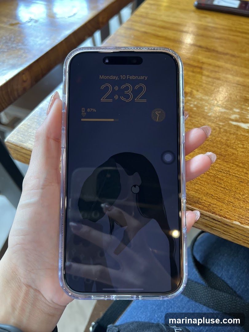 A person is holding an iPhone with the lock screen displayed. The time is 2:32 on Monday, 10 February. The battery is at 87%. There is a circular icon with the word "CUP" and a graphic of a cup. The wallpaper is a dark silhouette of a person's head and shoulders. The phone is on a wooden table.