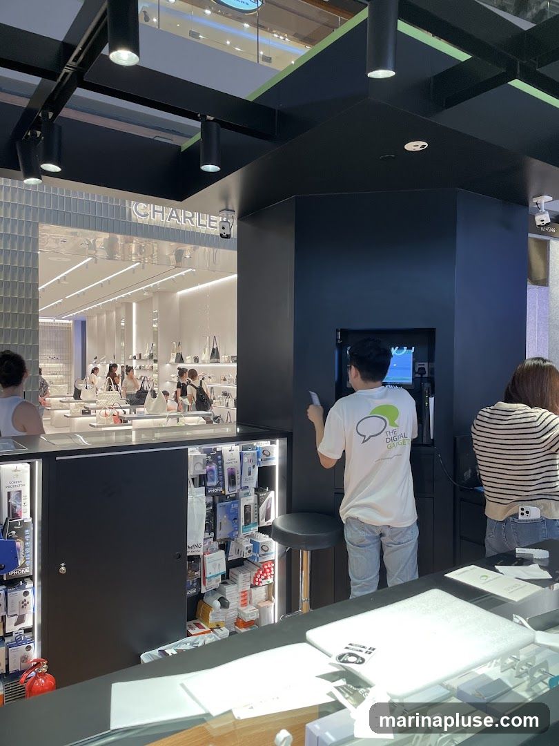 A view of a counter in an electronics store. A person in a white t-shirt with a logo that says "THE DIGITAL GADGET" is standing behind the counter, facing a screen. To the left, there are shelves stocked with phone accessories like cases and screen protectors. To the right, another person is standing. In the background, the store "CHARLES & KEITH" is visible, with displays of handbags and other accessories.