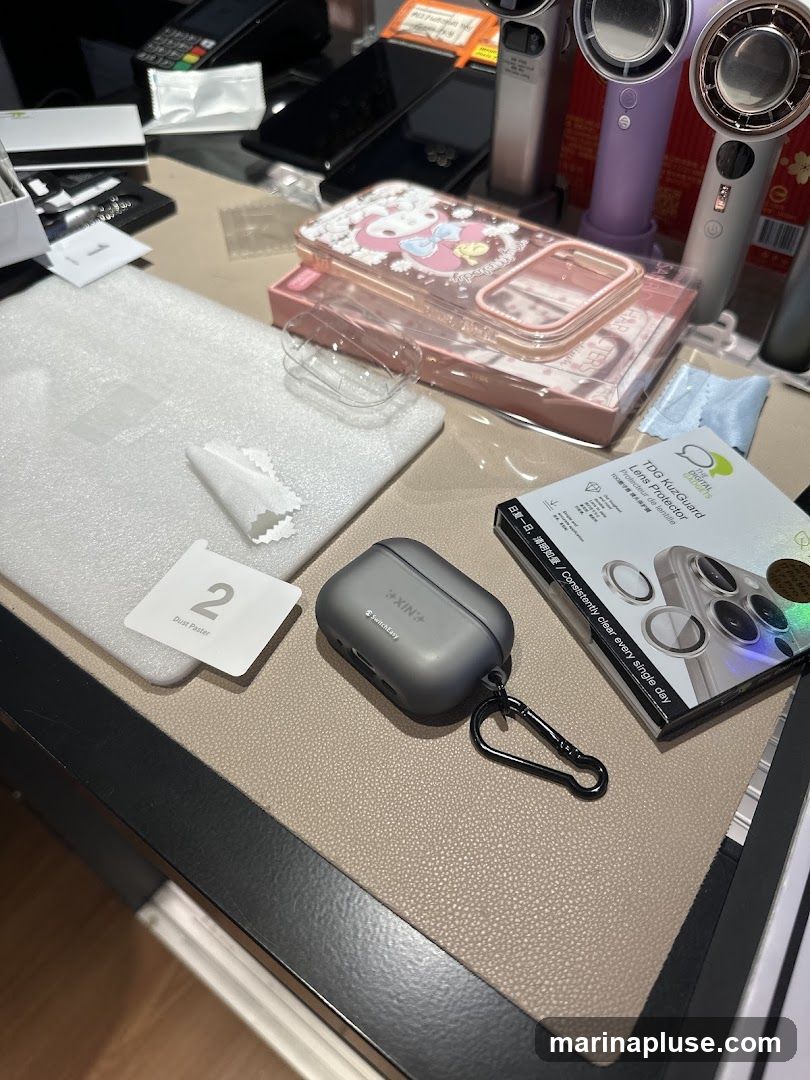 A collection of phone accessories including a dark grey AirPod case with a carabiner clip, a pink phone case with a My Melody design, a screen protector kit labeled 'TDG KuzGuard Lens Protector', and a 'Dust Paster' sticker. Several smartphones and other electronic devices are visible in the background.