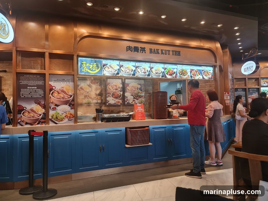 A food stall with a prominent sign reading "Bak Kut Teh" in English and Chinese. The stall displays various food items on a menu board with images and prices. Customers are seen ordering and waiting for their food. The stall has a blue cabinet base and a copper-colored counter and overhead structure.