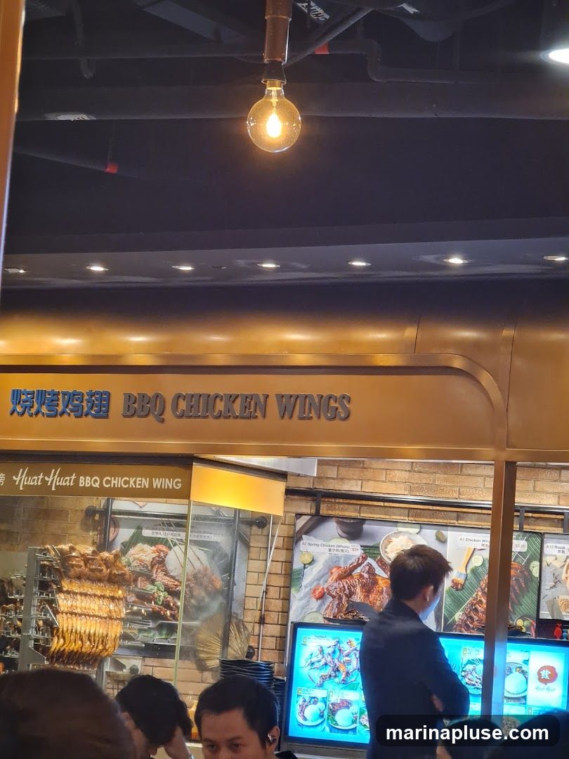A food stall with a prominent sign reading "BBQ CHICKEN WINGS" in English and Chinese characters. Below the main sign, smaller text reads "Huat Huat BBQ CHICKEN WING". The stall displays various images of grilled chicken and other dishes. A rack of golden-brown chicken wings is visible on the left. Several people are seen in the foreground, appearing to be customers. The interior features exposed pipes and a decorative light bulb hanging from the ceiling.