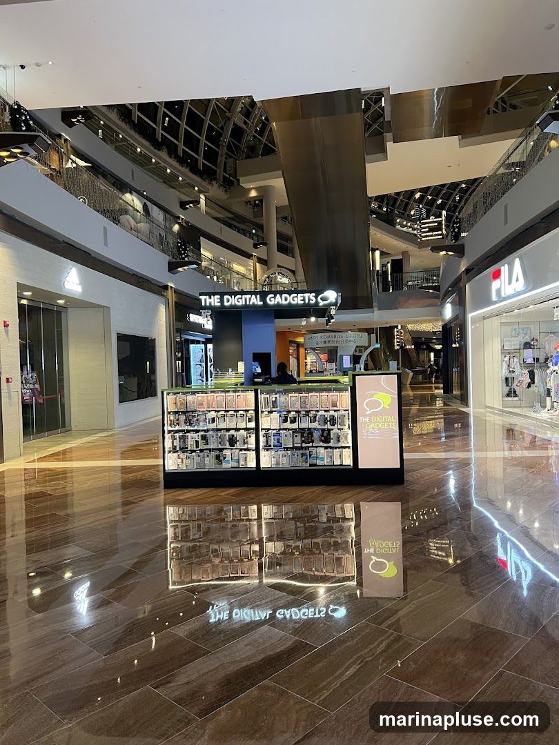 A kiosk in a shopping mall selling phone accessories. The kiosk has a display of phone cases and other electronic accessories. The signage above the kiosk reads "THE DIGITAL GADGETS". A promotional stand with the same branding is also visible. The floor is highly reflective, showing the reflection of the kiosk and surrounding lights.