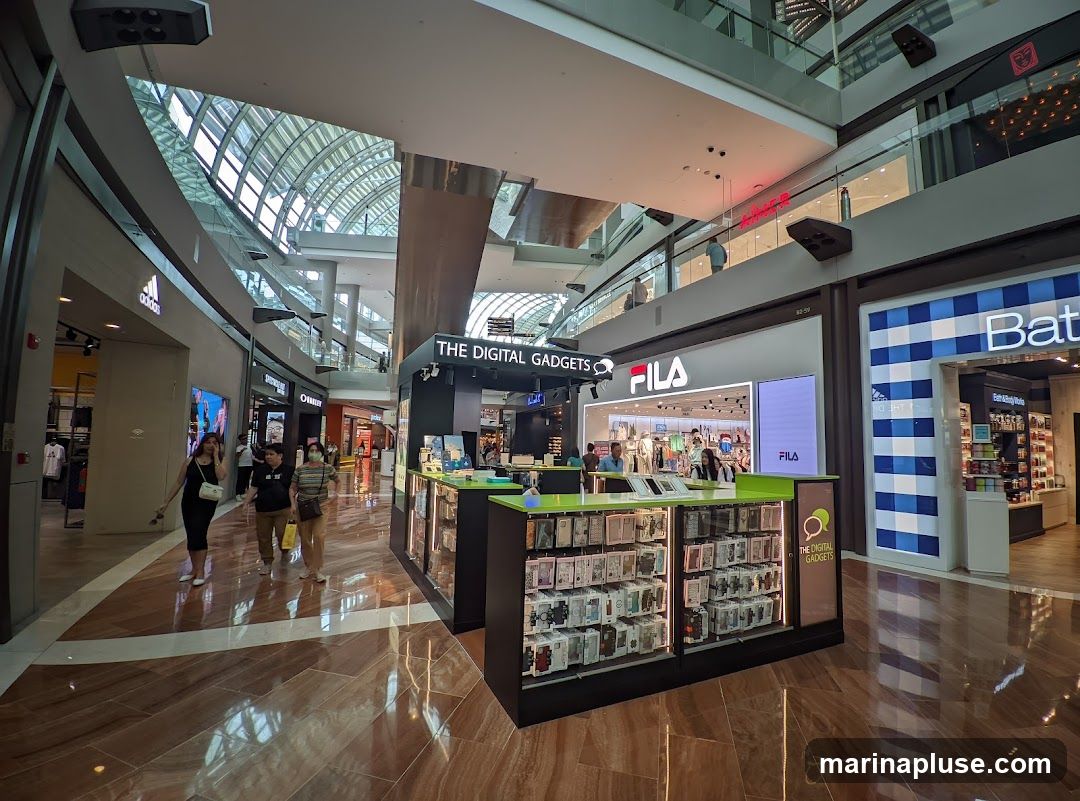A kiosk-style store named "THE DIGITAL GADGETS" is displayed in a shopping mall. The kiosk features numerous phone cases and accessories. In the background, other stores like Adidas, Fila, and Bath & Body Works are visible. People are walking around the mall.