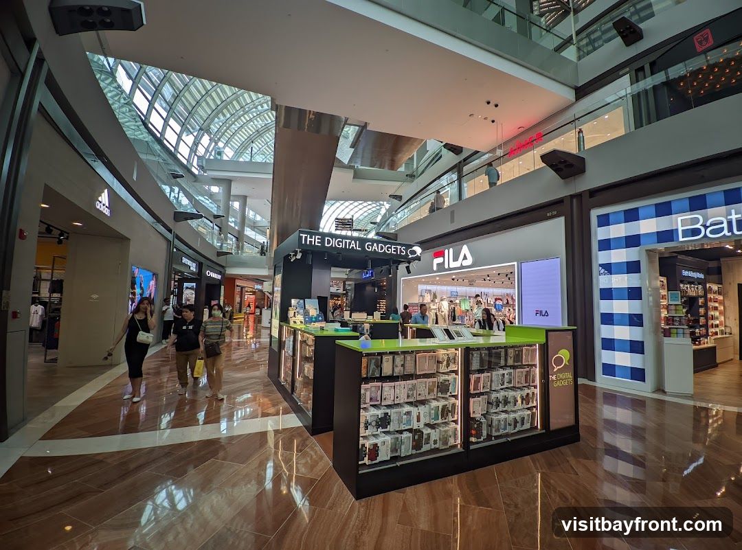 A kiosk-style store named "THE DIGITAL GADGETS" is displayed in a shopping mall. The kiosk features numerous phone cases and accessories. In the background, other stores like Adidas, Fila, and Bath & Body Works are visible. People are walking around the mall.
