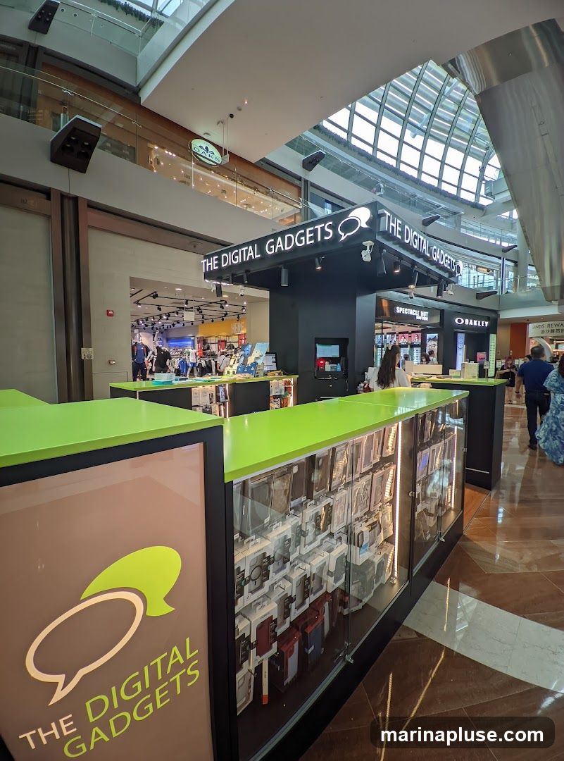 A kiosk-style store named "The Digital Gadgets" is displayed in a mall. The store features a black structure with illuminated signage and a bright green countertop. Display cases showcase a variety of phone cases and accessories. In the background, other stores like "Spectacle Hut" and "Oakley" are visible, along with shoppers.