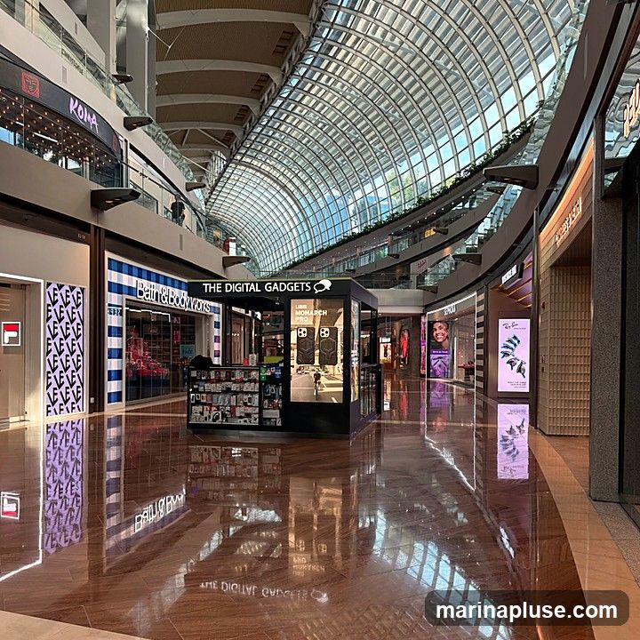 A wide shot of a modern shopping mall interior with a curved glass ceiling. Various shops are visible along the sides, including Bath & Body Works and a kiosk labeled "THE DIGITAL GADGETS". The polished wooden floor reflects the shops and the ceiling.