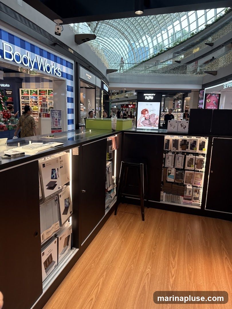 A view of a kiosk in a shopping mall selling electronic accessories. The kiosk has a black counter with various phone cases and tablet covers displayed. To the left, the storefront of 'Body Works' is visible with holiday-themed decorations. To the right, a display for 'ASTROCITY' features a promotional image of a person. The mall's interior with its distinctive arched ceiling and upper levels is visible in the background.