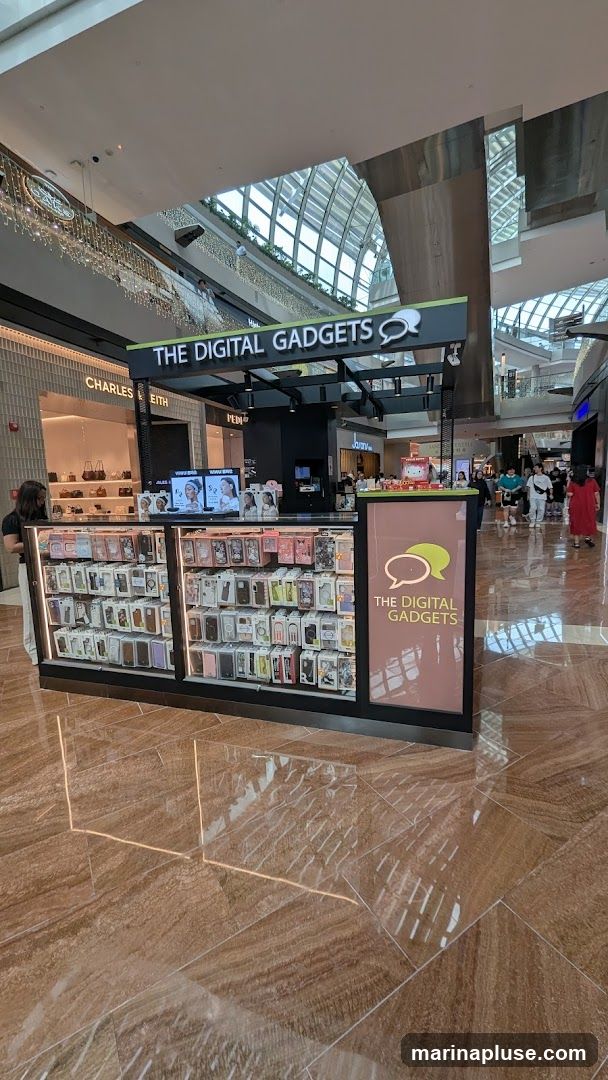 A kiosk in a shopping mall selling phone cases and other digital accessories. The kiosk has a prominent sign that says "THE DIGITAL GADGETS" with a speech bubble logo. The front of the kiosk displays a wide variety of phone cases organized by brand and model. In the background, other shops and shoppers are visible.