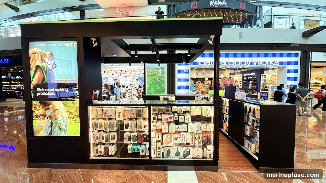 A kiosk in a mall selling phone cases and accessories. The kiosk has large display screens showing advertisements for phone cases. The display cases are filled with various phone accessories, including headphones and chargers. In the background, other stores are visible, including Bath & Body Works.