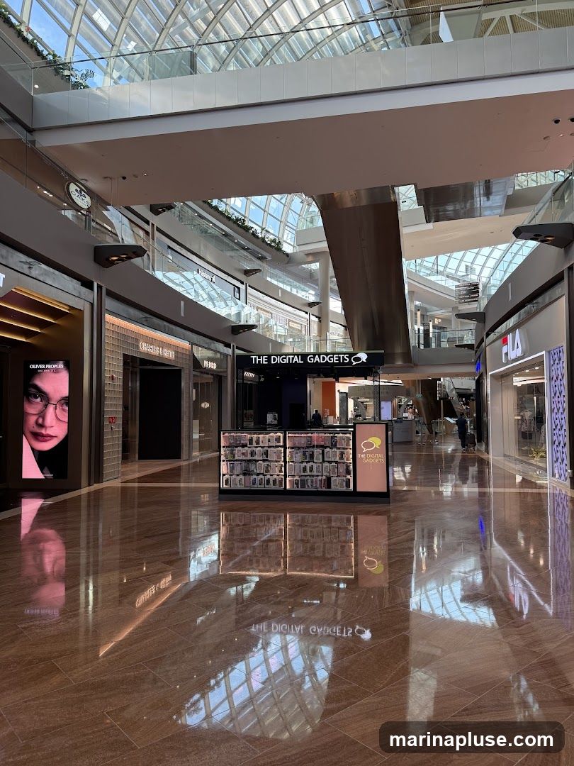A kiosk labeled "THE DIGITAL GADGETS" is centrally located in a modern, well-lit shopping mall. The kiosk displays various phone cases and accessories. In the background, other storefronts like Fila and Oliver Peoples are visible, along with the mall's architecture featuring a glass ceiling and multiple levels. The polished floor reflects the overhead lights and structures.