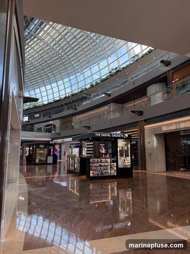 A view of a shopping mall interior with a central kiosk selling digital gadgets. The kiosk has a sign that reads "THE DIGITAL GADGETS". Various electronic accessories are displayed on shelves and in cases. In the background, other stores like Sephora and Charles & Keith are visible. The mall features a large, arched glass ceiling that allows natural light to illuminate the space. The floors are polished and reflective.