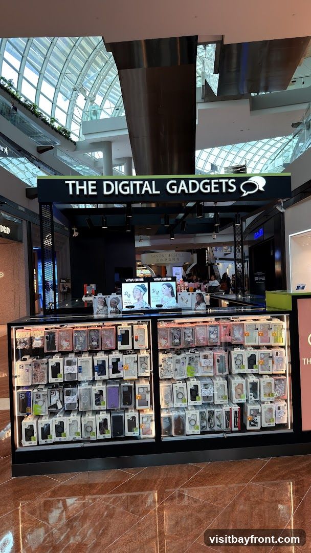 A kiosk in a mall selling phone cases and other digital accessories. The kiosk has a prominent sign that reads "THE DIGITAL GADGETS" with a speech bubble logo. The display shelves are filled with various phone cases, organized by brand and design. In the background, there are other shops and mall architecture visible.