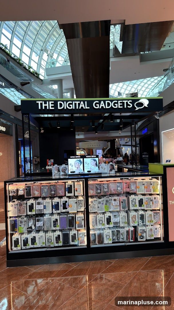 A kiosk in a mall selling phone cases and other digital accessories. The kiosk has a prominent sign that reads "THE DIGITAL GADGETS" with a speech bubble logo. The display shelves are filled with various phone cases, organized by brand and design. In the background, there are other shops and mall architecture visible.