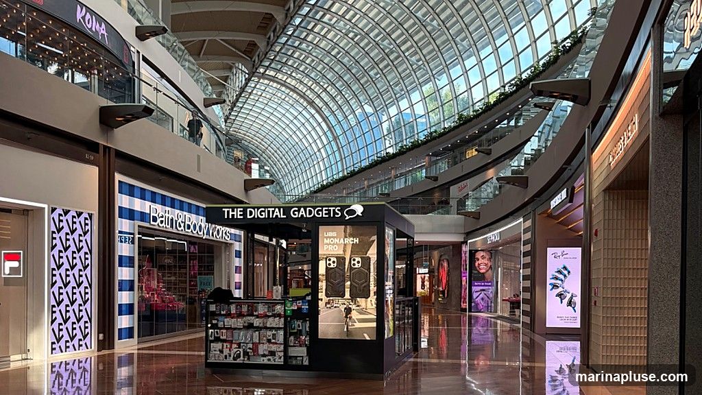 An interior view of a modern shopping mall with a distinctive glass-domed ceiling. Several storefronts are visible, including Bath & Body Works, The Digital Gadgets kiosk, Sephora, and a Ray-Ban display. The mall features multiple levels with escalators and walkways. The flooring is highly reflective, mirroring the architecture and lighting above.