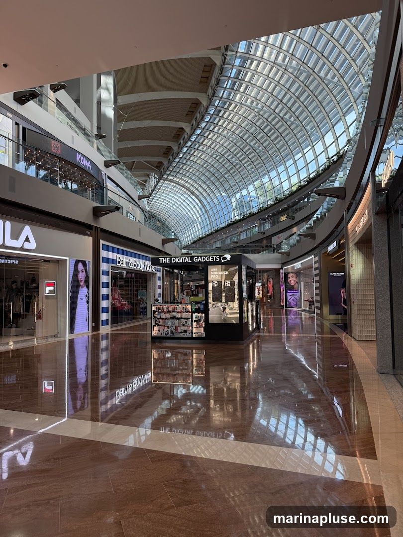 A wide shot of a modern shopping mall interior with a distinctive glass-paneled, arched ceiling. Various storefronts are visible along the sides, including Fila and Bath & Body Works. In the center, a kiosk labeled "THE DIGITAL GADGETS" displays electronic accessories. The polished floor reflects the architecture and lighting of the mall.
