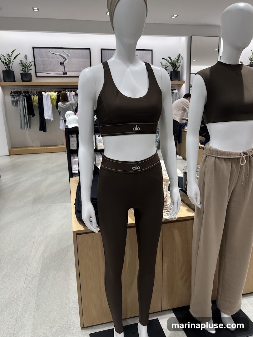 Mannequins are displayed wearing athletic wear in a store. The mannequin in the foreground wears a brown sports bra and matching leggings, both with the 'alo' logo. Another mannequin to the right wears a brown sleeveless top and tan sweatpants. In the background, clothing racks with various apparel are visible, along with artwork on the wall and plants.