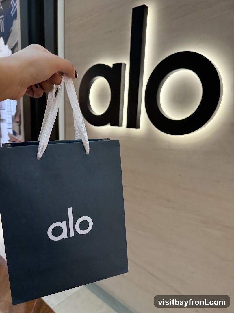 A hand holds a dark blue shopping bag with the white logo "alo" printed on it. In the background, a large, illuminated black "alo" logo is mounted on a textured wall.
