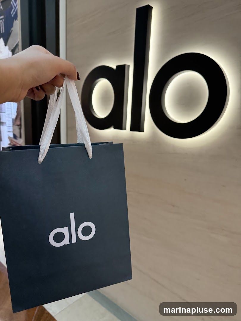 A hand holds a dark blue shopping bag with the white logo "alo" printed on it. In the background, a large, illuminated black "alo" logo is mounted on a textured wall.