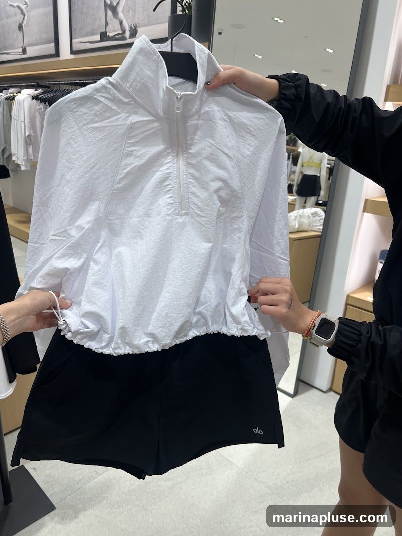 A person is holding up a white, quarter-zip pullover with a high collar and cinched waist, paired with black athletic shorts. The shorts have a small logo on the lower right leg. In the background, there are racks of clothing and framed artwork on the wall, suggesting a retail environment.