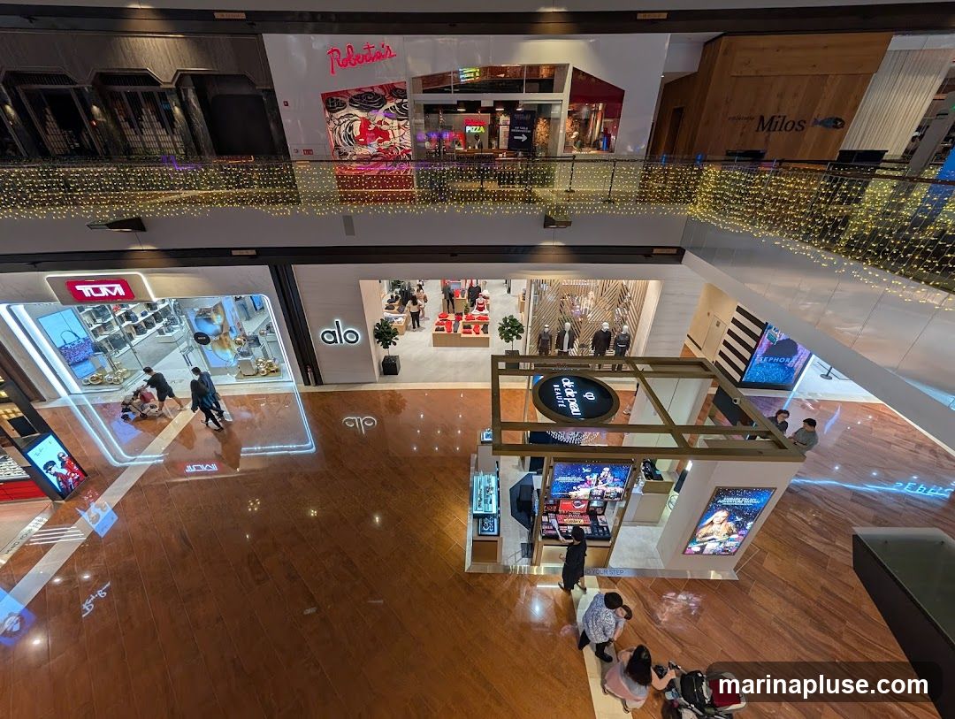 An overhead view of a modern shopping mall interior. On the upper level, storefronts like 'Roberts' and 'Pizza' are visible, adorned with festive string lights. The lower level features various retail spaces, including 'TUM', 'alo', and a 'Milos' restaurant. A prominent kiosk in the center displays beauty products, with shoppers gathered around. The polished wooden floor reflects the ambient lighting and store displays.