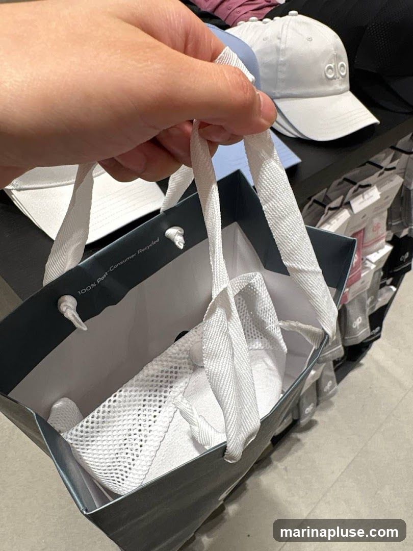 A hand holds a shopping bag containing white sneakers. The bag is dark gray with white handles and has text that reads "100% Post-Consumer Recycled." In the background, shelves are stocked with white baseball caps, some with the "alo" logo visible, and other apparel.