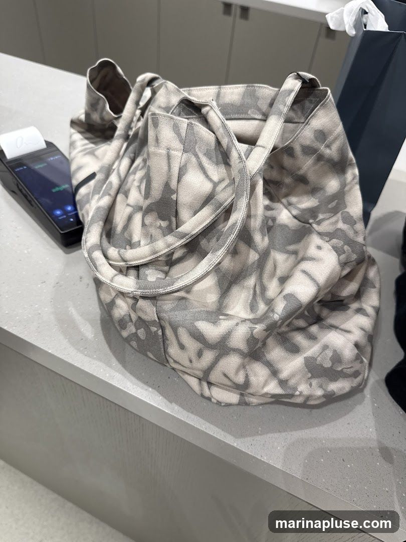 A tie-dye patterned tote bag rests on a counter next to a payment terminal. The bag is made of a light-colored fabric with a grey tie-dye pattern. The payment terminal is black and has a small screen displaying some text and buttons. In the background, a dark blue shopping bag is visible.