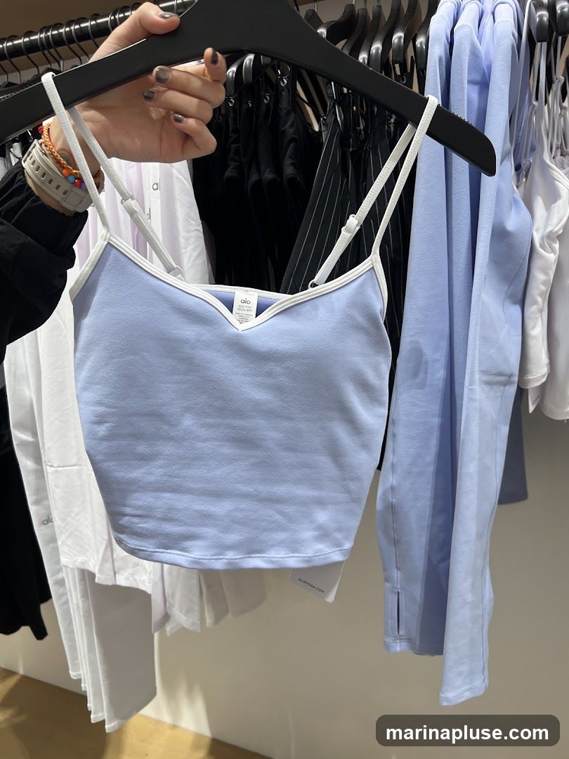 A light blue cropped tank top with white trim is displayed on a hanger. The tank top has thin white straps and a V-neckline. Behind it, other clothing items, including white tops and black garments, are visible on hangers in a store setting. A tag with the logo "alo" is visible on the tank top.