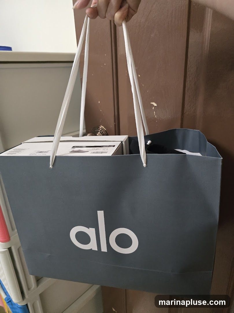 A dark gray shopping bag with white handles and the white logo 'alo' is being held by a person. Inside the bag, boxes and other items are visible. The bag appears to be made of paper.
