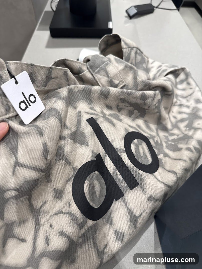 A close-up shot of a tote bag with a tie-dye pattern and the "alo" logo printed in black. A white tag with the "alo" logo is attached to the bag. The bag is placed on a countertop.