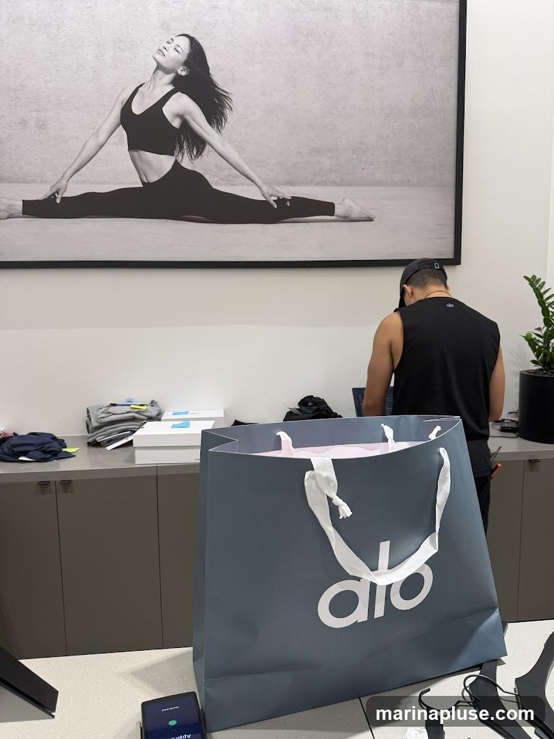 A large gray shopping bag with a white logo is prominently displayed in the foreground. Behind it, a person wearing a black tank top and a baseball cap is seen from the back, working at a counter with a laptop. The wall behind them features a large black and white photograph of a woman in a yoga pose. The counter area shows folded clothes and boxes.