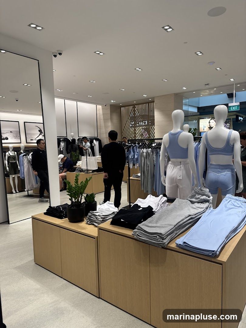 The image shows the interior of a modern clothing store. Racks of clothes are visible, along with mannequins displaying athletic wear. Folded stacks of t-shirts and sweatpants in various colors (black, grey, light blue) are neatly arranged on wooden display units. A large mirror is on the left side of the frame. Several customers and staff members are present in the store. An "EXIT" sign is visible in the background.
