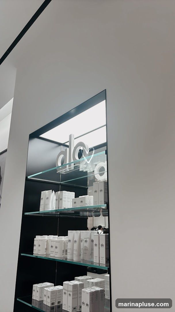 A mirrored display shelf showcases various Alo branded skincare products. The products are predominantly white with the Alo logo visible. The shelves are made of glass and are illuminated from above.