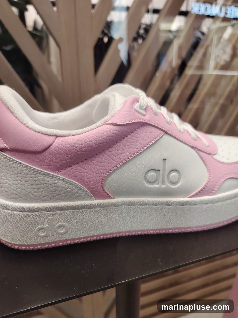 A close-up view of a pink and white sneaker with the "alo" logo embossed on the side and the sole. The sneaker has white laces and a white sole with a pink trim. The background is blurred, showing a retail display with wooden elements and some clothing racks.