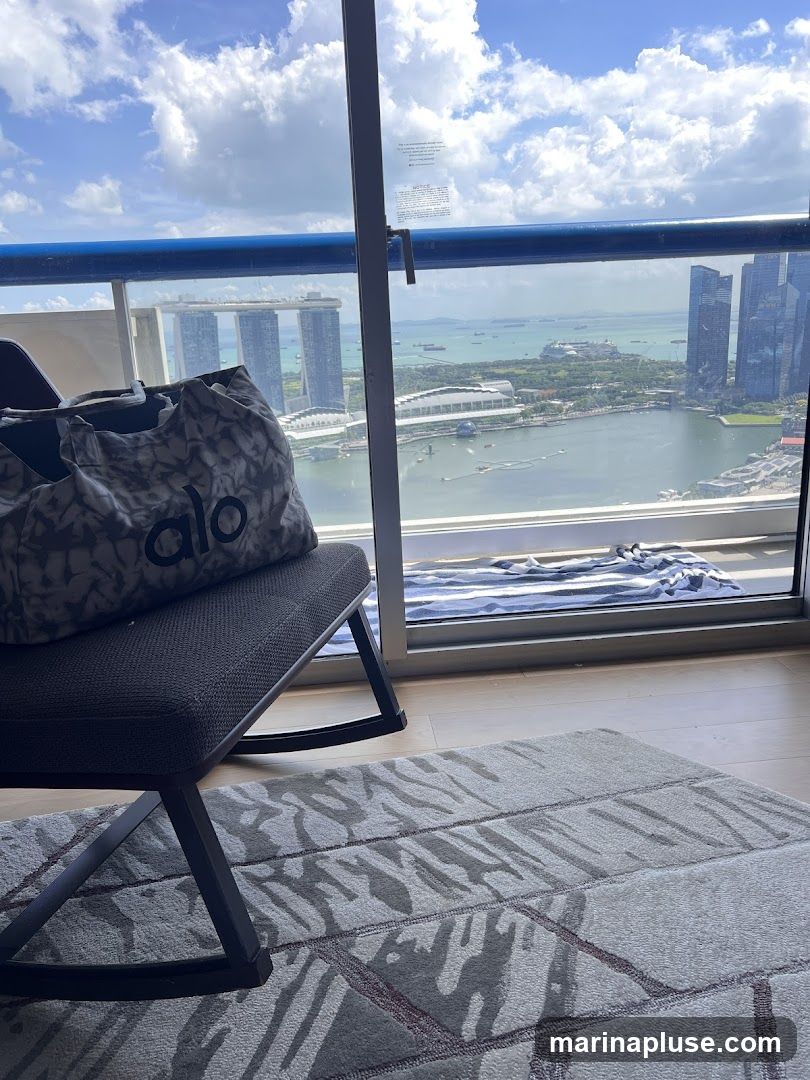 A tote bag with the 'alo' logo rests on a rocking chair positioned on a balcony. The bag is grey with a tie-dye pattern. The rocking chair is dark grey. In the background, a panoramic view of a city skyline is visible through a large window, featuring prominent skyscrapers and a body of water with boats. The sky is blue with scattered white clouds. A patterned rug is on the floor in the foreground.