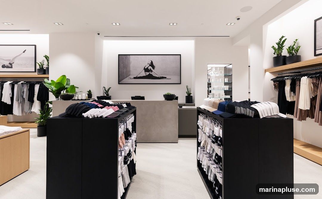 A bright and modern retail store interior featuring clothing racks with apparel, display shelves with hats and socks, and a central counter. Black display units showcase folded hats and packaged socks. Racks on the left display various tops and bottoms in neutral colors. A large framed photograph of a woman in a yoga pose hangs above the counter. Plants are strategically placed throughout the store to add a touch of nature. A glass display case with products is visible in the background.