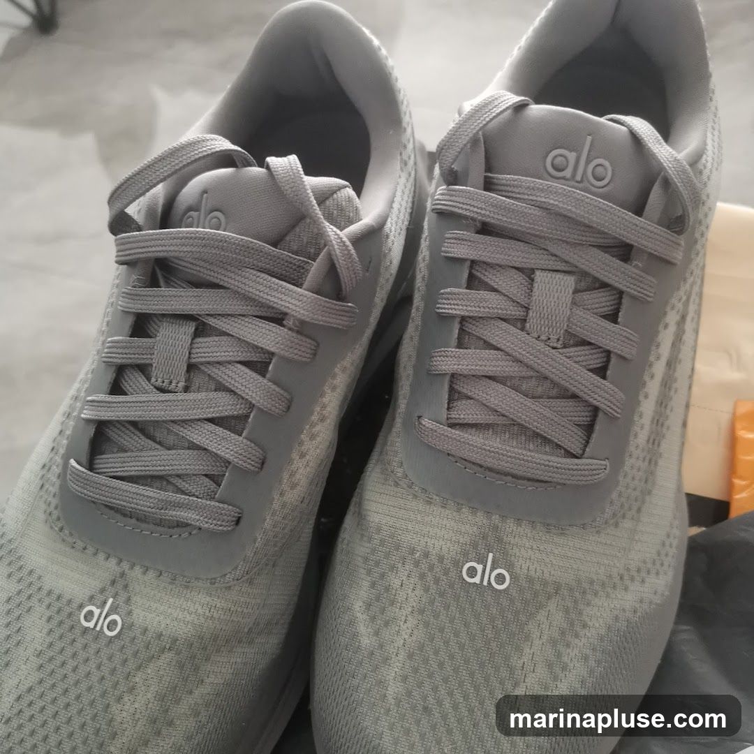A pair of gray Alo sneakers with white logos on the tongue and toe box. The shoes are laced and appear to be new, possibly in packaging.