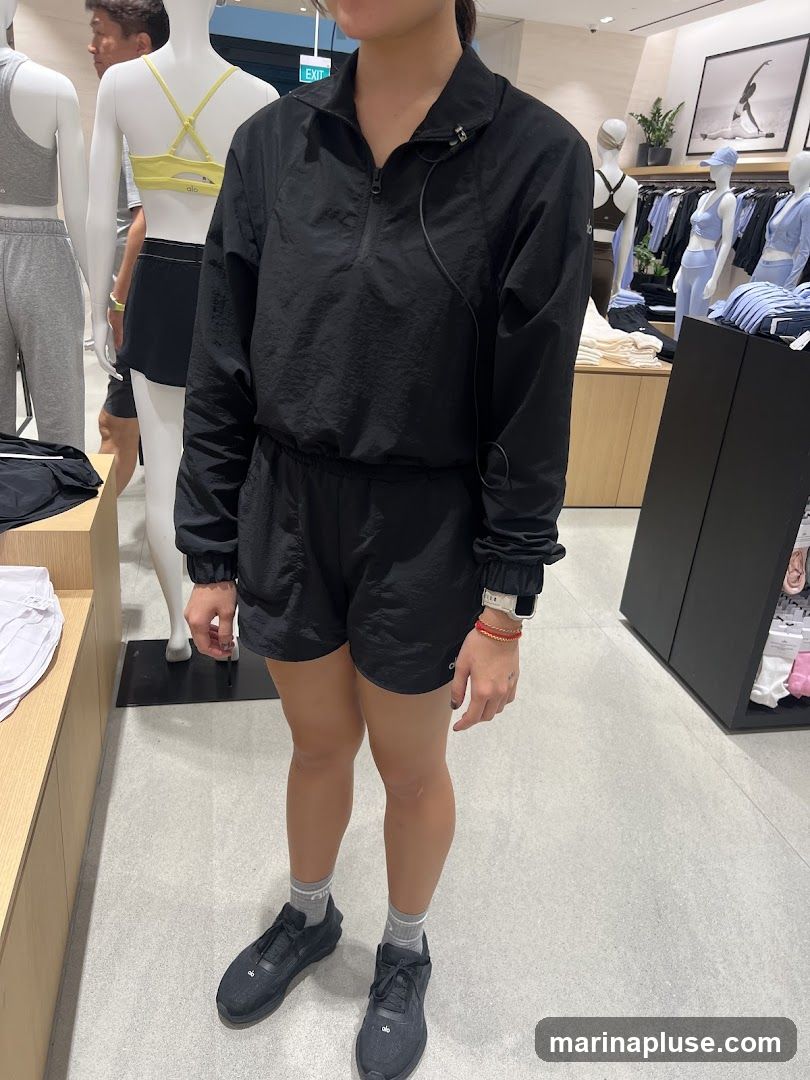 A woman stands in a retail store, wearing a black half-zip jacket and matching shorts. She has on black athletic shoes and grey socks. In the background, mannequins display athletic apparel, including sports bras and leggings. The store appears to be a clothing boutique, with racks of clothing and display shelves visible.