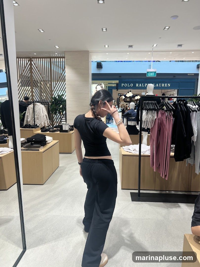A young woman with her back to the camera is posing with a peace sign in a well-lit clothing store. She is wearing a black crop top and black sweatpants. The store features wooden displays with folded clothing and racks of garments, including shirts and pants. In the background, a sign for "POLO RALPH LAUREN" is visible above an exit.