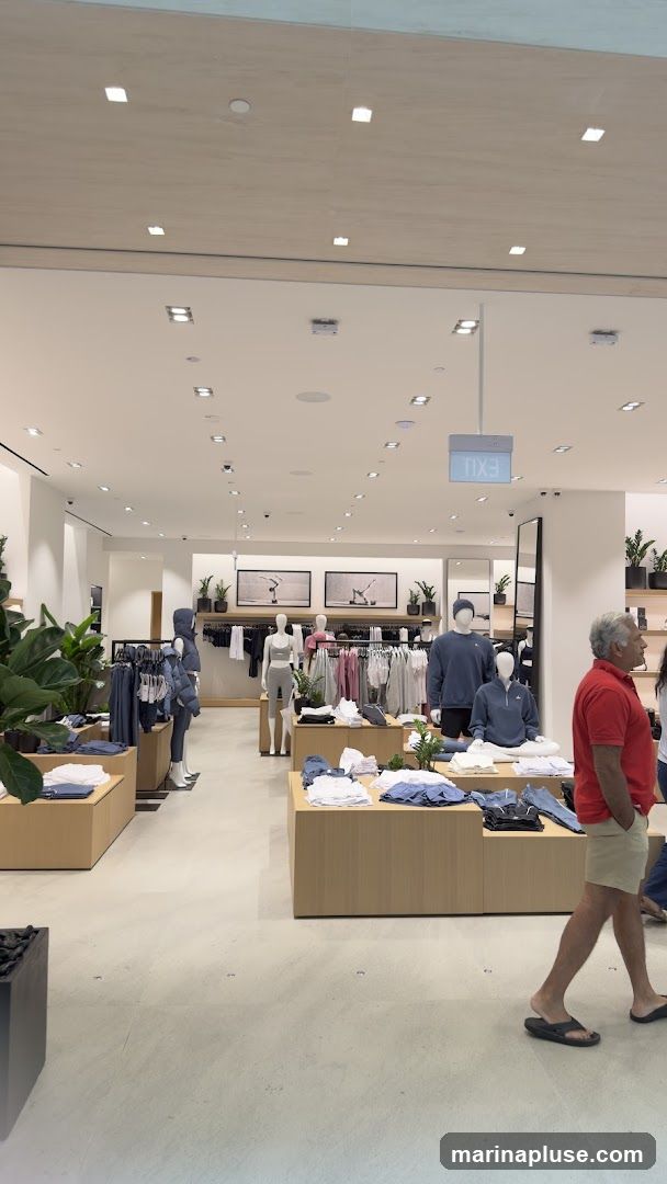 The image shows the interior of a modern clothing store. Mannequins are dressed in athletic wear, and racks of clothing are neatly displayed on wooden shelves and hanging racks. The store features a clean, minimalist design with bright lighting and potted plants. An illuminated 'EXIT' sign is visible hanging from the ceiling. A man in a red shirt and khaki shorts is walking through the store.