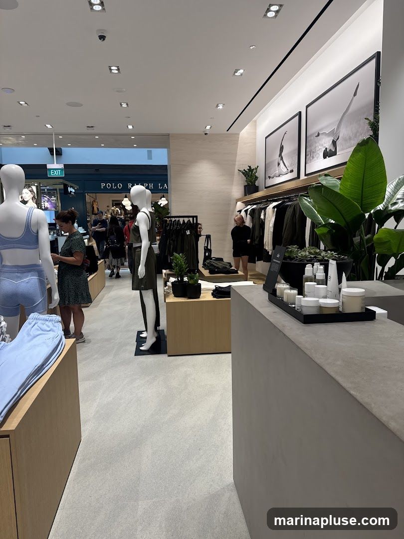 The image shows the interior of a modern clothing store. In the foreground, a counter displays various skincare products. To the left, mannequins are dressed in athletic wear, with a display of light blue sweatpants. Further into the store, racks of clothing are visible, and in the background, a sign for 'POLO RALPH LAUREN' is displayed. The store features minimalist decor with large framed artwork on the walls and lush green plants.
