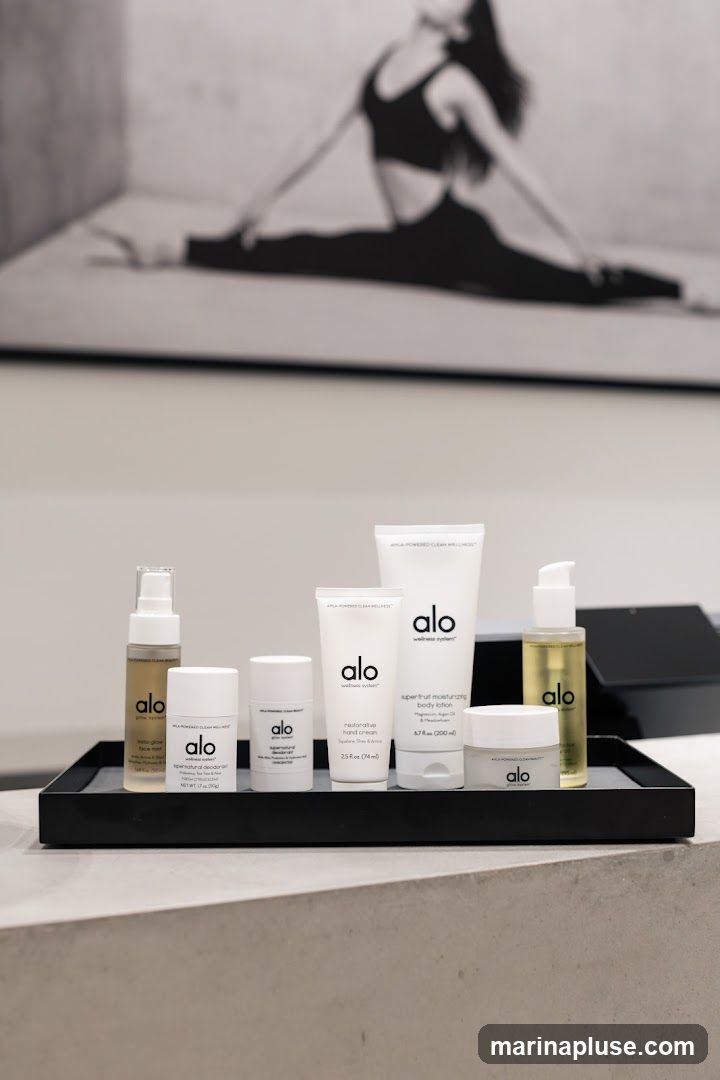 A black tray holds several Alo wellness products, including a face mist, deodorant, hand cream, body lotion, and a jar of cream. A black and white photograph of a woman in a yoga pose is visible in the background.