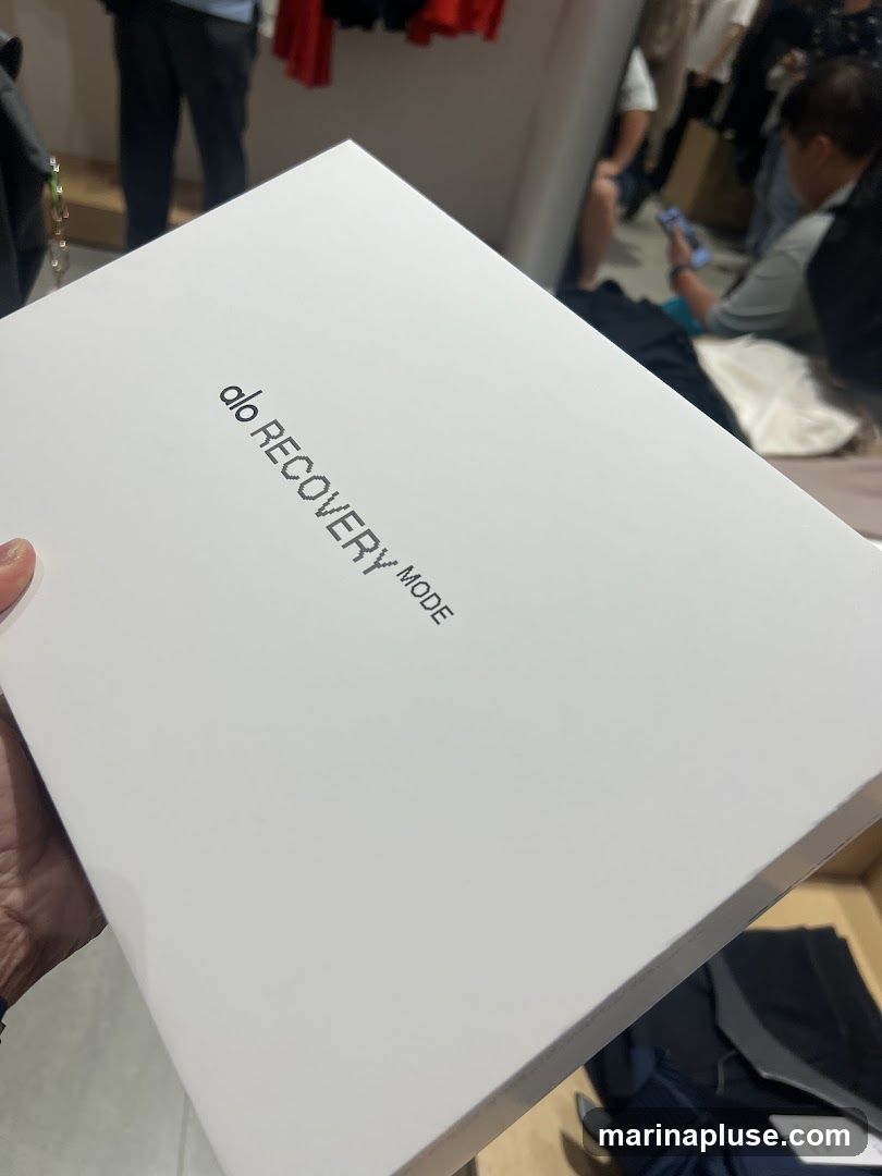A white box with the text "alo RECOVERY MODE" printed on it. The box is held by a hand. In the background, there are blurred figures of people and clothing racks, suggesting an indoor retail environment.