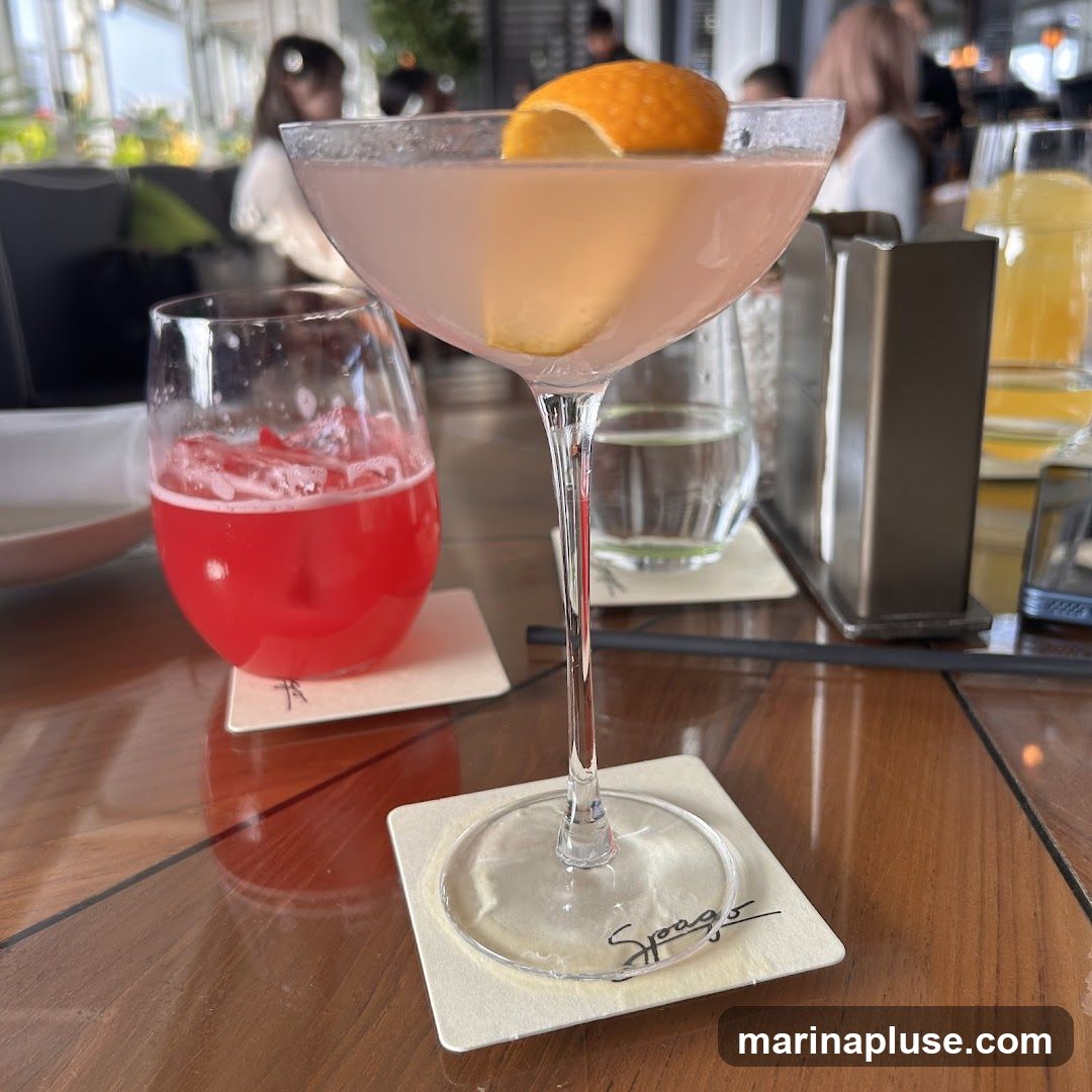 Two cocktails are on a wooden table. The cocktail in the foreground is in a coupe glass and is a pale pink color, garnished with an orange peel. The cocktail in the background is in a rocks glass and is a vibrant red color. Other drinks and people are visible in the blurred background.